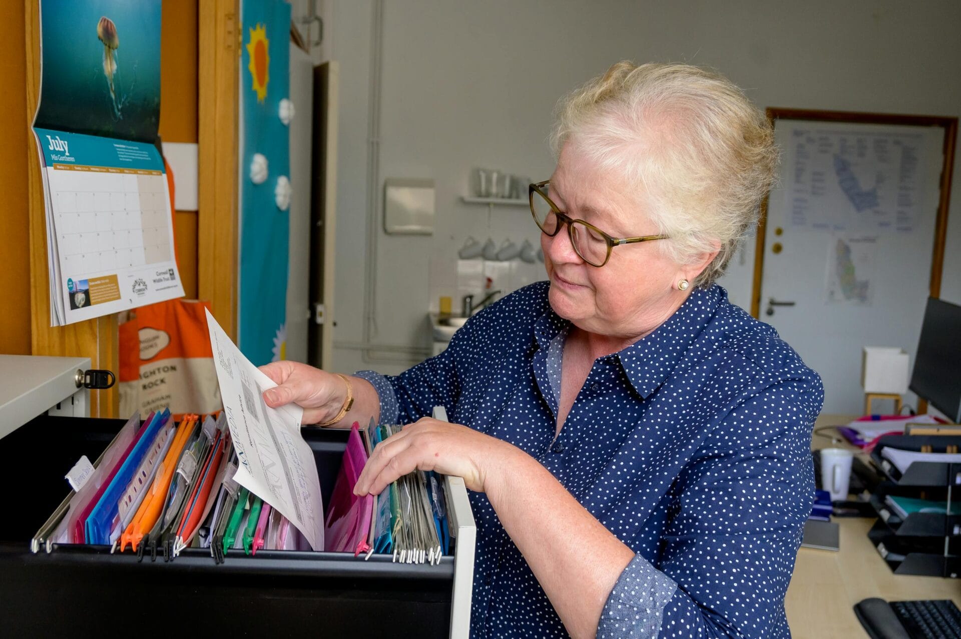 Elderly woman sorting paperwork in an office setting with organized files.