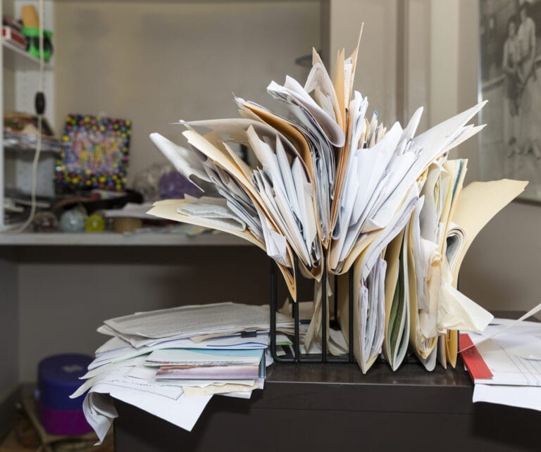 NAID AAA Certification: desk clutter with haphazardly arranged, overstuffed file folders in a rack on a messy desk in a cluttered room